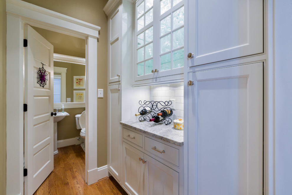 Example of a mid-sized transitional l-shaped light wood floor kitchen design in Cincinnati with a farmhouse sink, beaded inset cabinets, white cabinets, granite countertops, white backsplash, porcelain backsplash, white appliances and an island
