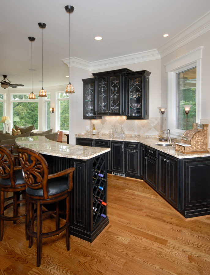 Photo of a large traditional u-shaped seated home bar in Cincinnati with an undermount sink, raised-panel cabinets, black cabinets, granite benchtops, beige splashback, stone tile splashback and medium hardwood floors.