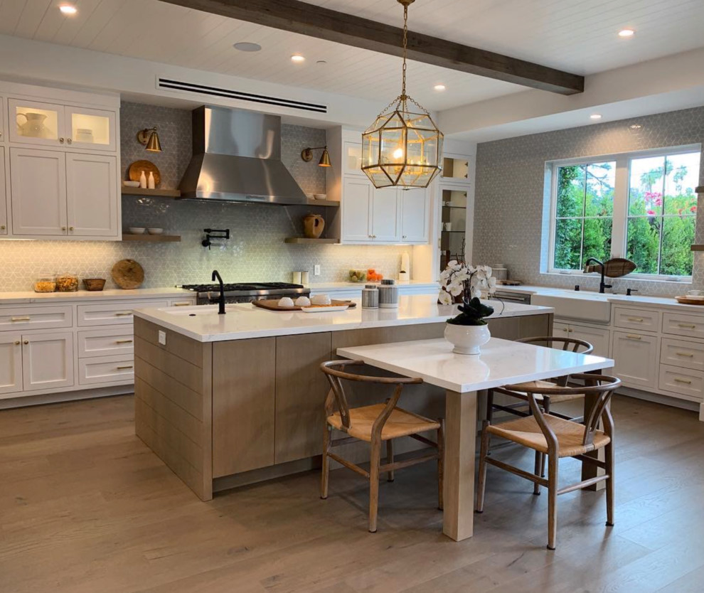 Design ideas for a large country l-shaped open plan kitchen in Los Angeles with glass-front cabinets, white splashback, ceramic splashback, stainless steel appliances, light hardwood floors, with island, white benchtop, a farmhouse sink, white cabinets, quartzite benchtops, brown floor, exposed beam and timber.