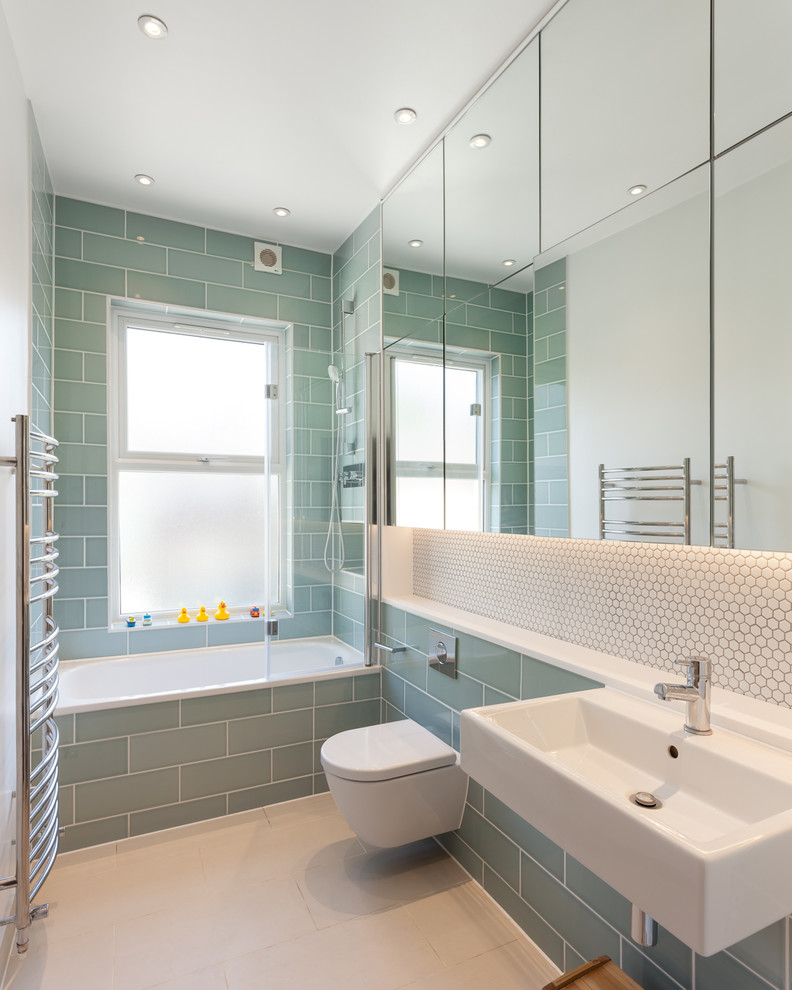 Inspiration for a transitional bathroom in London with flat-panel cabinets, a drop-in tub, a shower/bathtub combo, a wall-mount toilet, green tile, ceramic tile, ceramic floors, a wall-mount sink, beige floor and a hinged shower door.