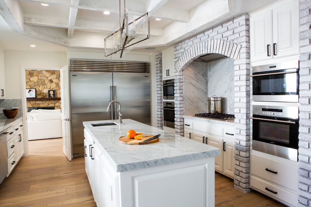 This is an example of a large beach style kitchen in Los Angeles with flat-panel cabinets, white cabinets, marble benchtops, white splashback, marble splashback, stainless steel appliances, light hardwood floors, with island, brown floor and white benchtop.