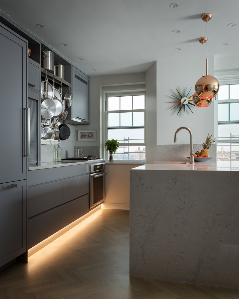 Contemporary galley kitchen in New York with a farmhouse sink, flat-panel cabinets, grey cabinets, grey splashback, stainless steel appliances, light hardwood floors, a peninsula, brown floor and white benchtop.