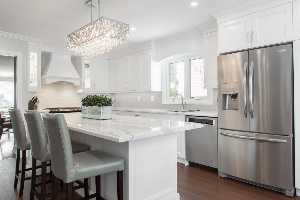 Large transitional l-shaped brown floor and dark wood floor enclosed kitchen photo with an undermount sink, recessed-panel cabinets, white cabinets, quartz countertops, gray backsplash, stainless steel appliances, an island, white countertops and subway tile backsplash
