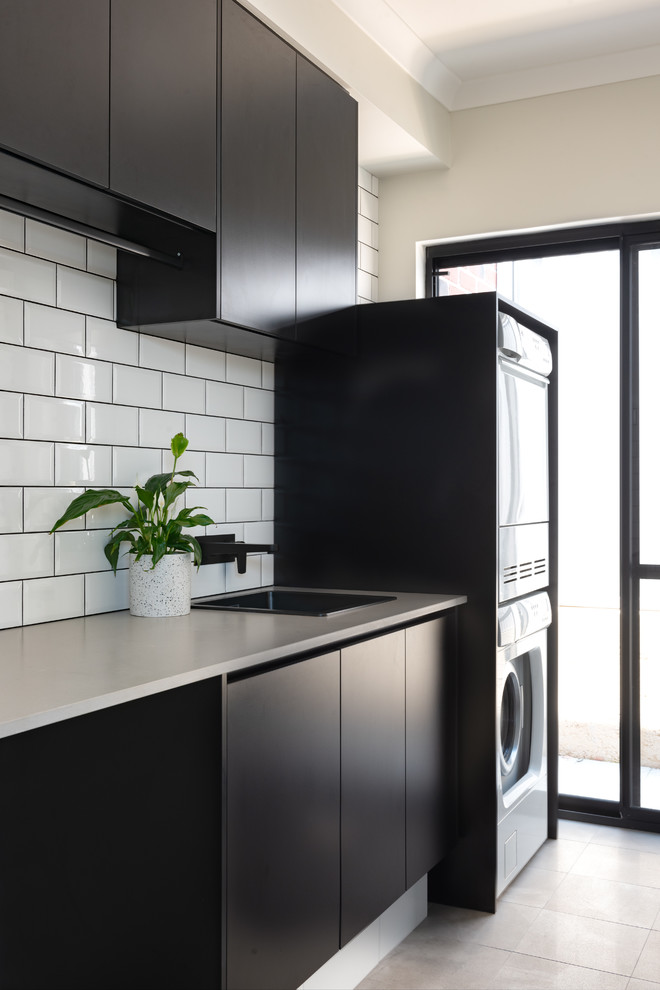 Industrial dedicated laundry room in Perth with a drop-in sink, black cabinets, quartz benchtops, ceramic floors, a stacked washer and dryer, grey floor and grey benchtop.