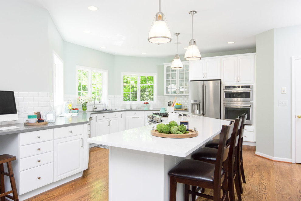 Example of a large transitional u-shaped medium tone wood floor and brown floor eat-in kitchen design in Detroit with an undermount sink, raised-panel cabinets, white cabinets, quartz countertops, white backsplash, ceramic backsplash, white appliances and an island