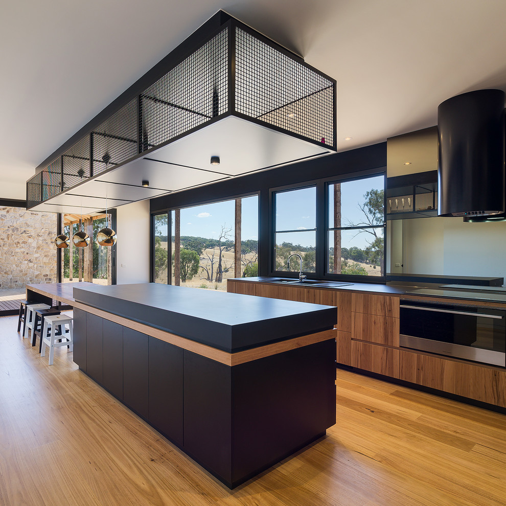Contemporary galley eat-in kitchen in Melbourne with with island, a drop-in sink, flat-panel cabinets, black cabinets, white splashback, stainless steel appliances, medium hardwood floors, brown floor and black benchtop.