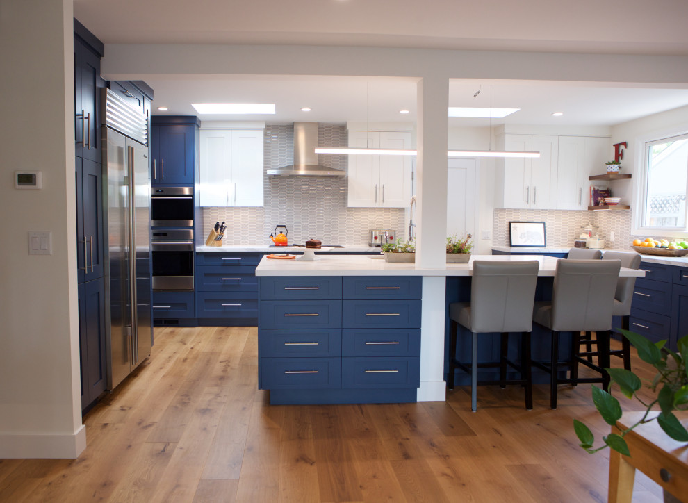 Large traditional u-shaped open plan kitchen in San Francisco with a single-bowl sink, recessed-panel cabinets, blue cabinets, engineered stone countertops, grey splashback, mosaic tiled splashback, stainless steel appliances, medium hardwood flooring, an island and white worktops.