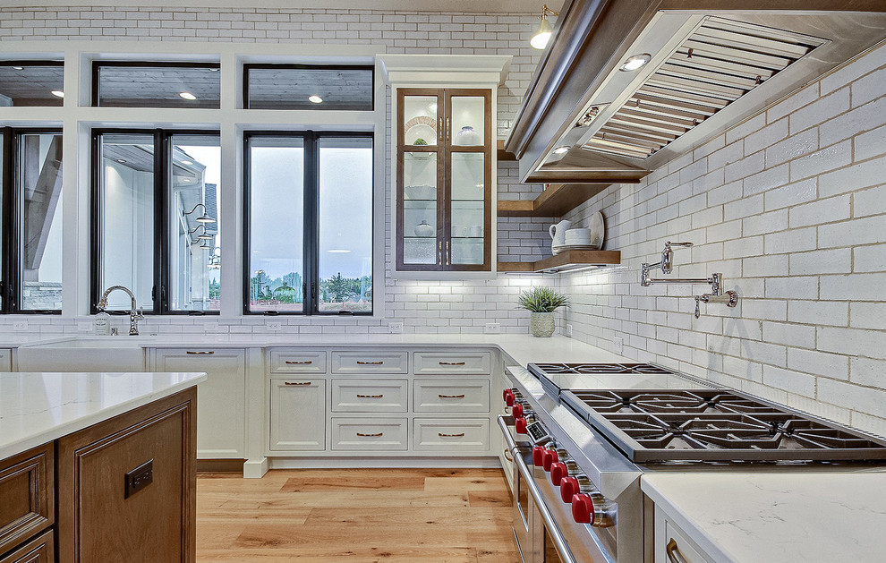 Design ideas for an expansive country u-shaped open plan kitchen in Portland with a farmhouse sink, recessed-panel cabinets, white cabinets, quartz benchtops, white splashback, ceramic splashback, stainless steel appliances, light hardwood floors, with island, brown floor and white benchtop.