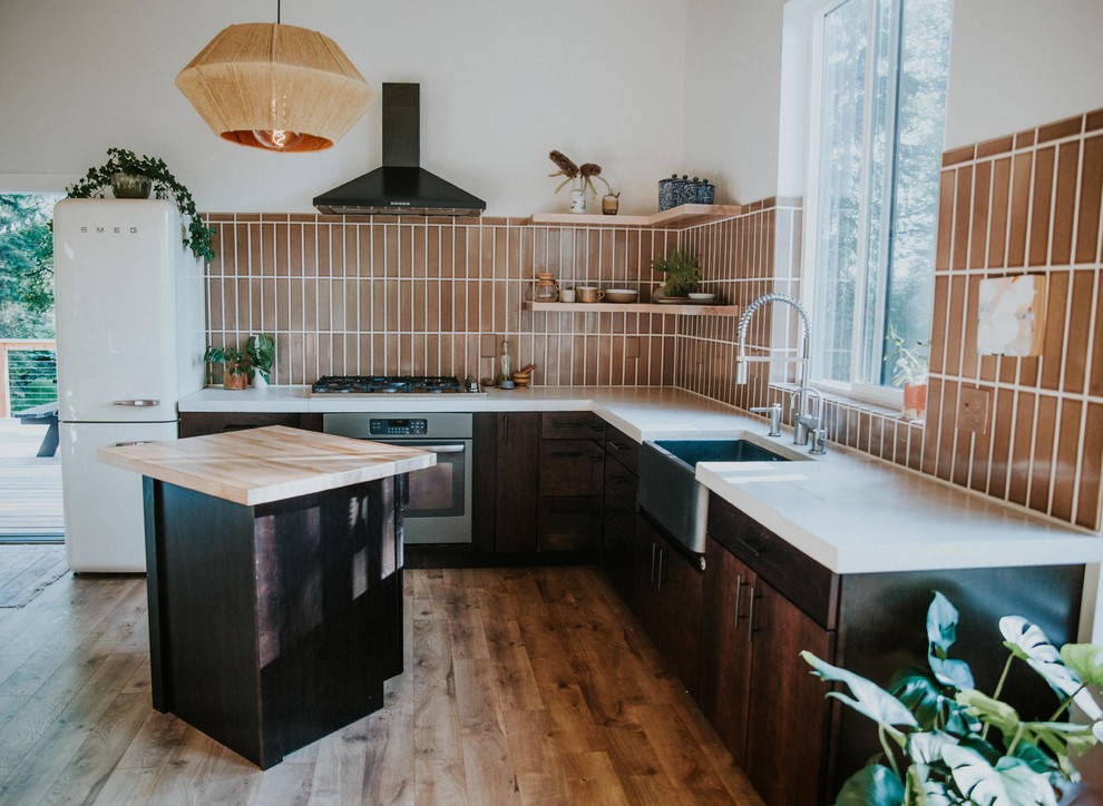 Photo of a mid-sized scandinavian l-shaped open plan kitchen in Other with dark wood cabinets, brown splashback, ceramic splashback, stainless steel appliances, medium hardwood floors, with island, brown floor and white benchtop.