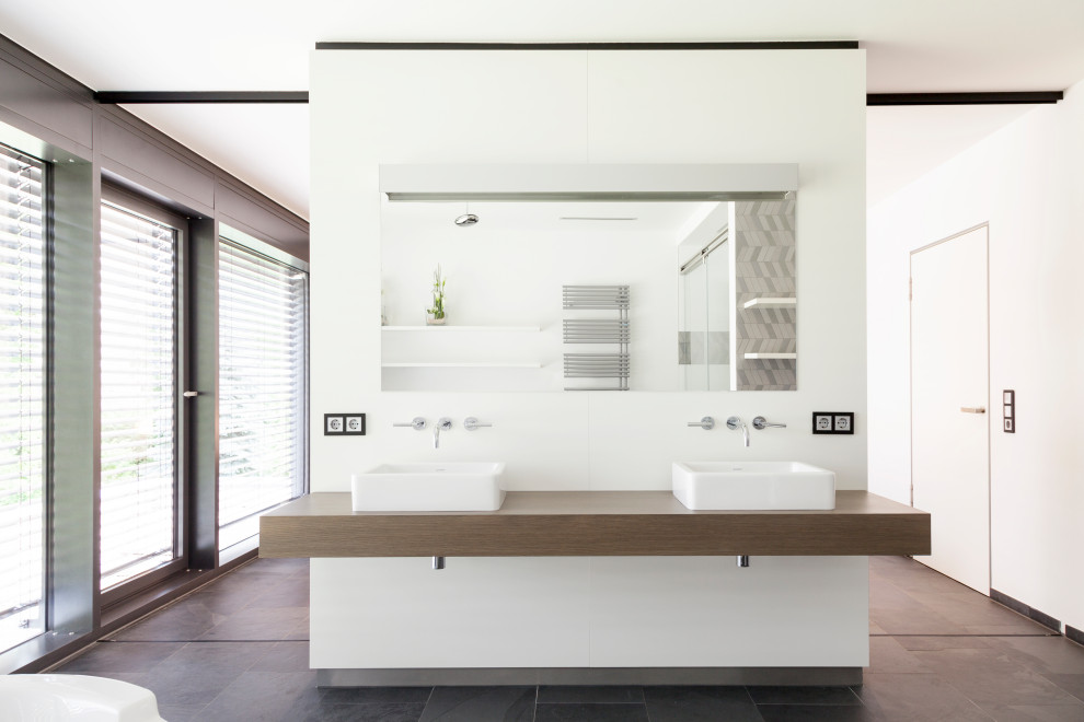 Photo of an expansive contemporary master bathroom in Other with flat-panel cabinets, dark wood cabinets, a freestanding tub, white tile, white walls, slate floors, a vessel sink, wood benchtops, grey floor, brown benchtops, a niche, a double vanity and a floating vanity.