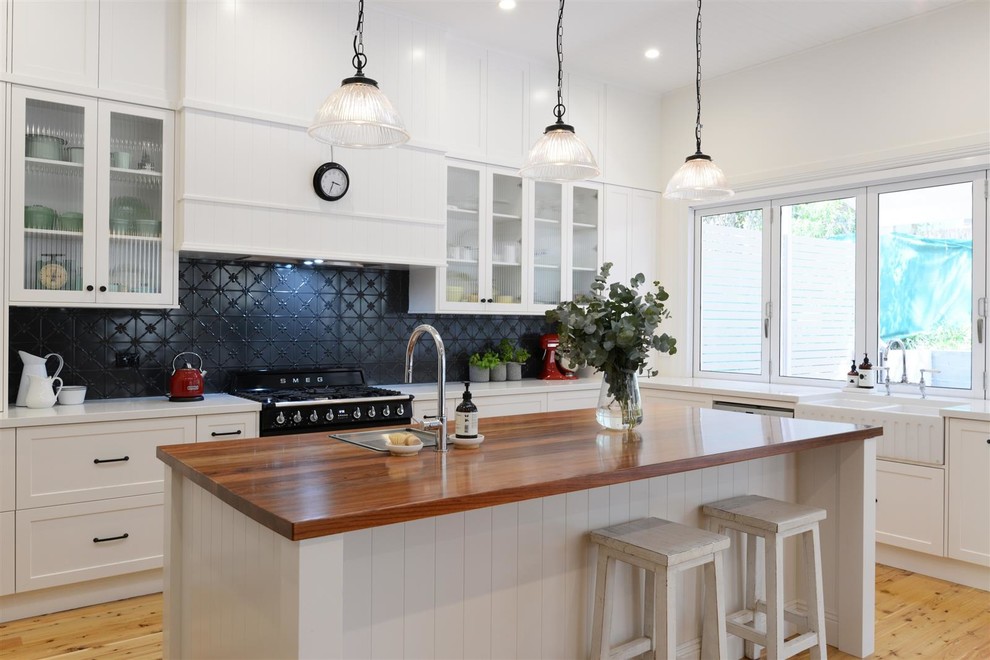 Photo of a large traditional u-shaped open plan kitchen in Sydney with a drop-in sink, open cabinets, white cabinets, wood benchtops, black splashback, ceramic splashback, black appliances, light hardwood floors, with island, brown floor and grey benchtop.