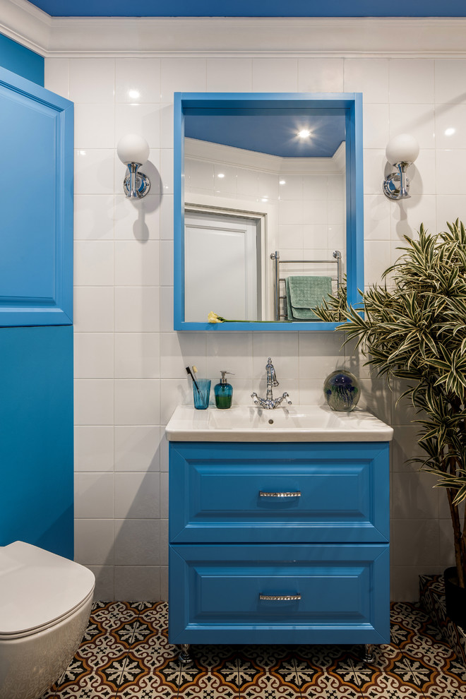 This is an example of a contemporary bathroom in Other with raised-panel cabinets, blue cabinets, a wall-mount toilet, white tile, an integrated sink and multi-coloured floor.