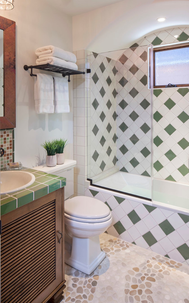 This is an example of a mediterranean bathroom in Los Angeles with an alcove tub, a shower/bathtub combo, green tile, a drop-in sink, louvered cabinets, medium wood cabinets, tile benchtops and green benchtops.