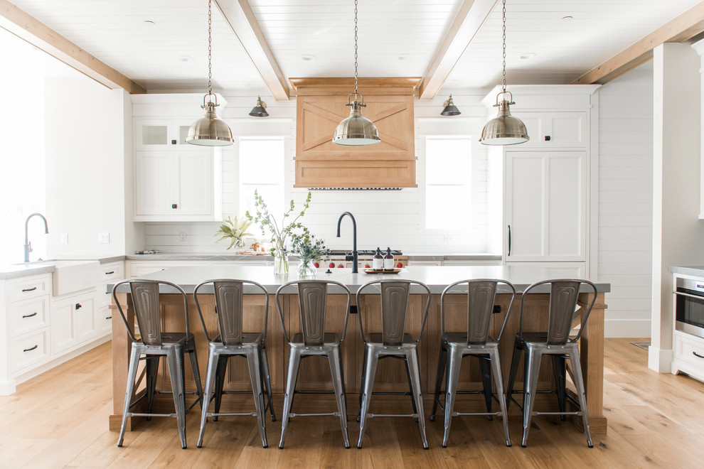Photo of a beach style l-shaped kitchen in Salt Lake City with shaker cabinets, white cabinets, white splashback, with island, grey benchtop, a farmhouse sink, stainless steel appliances, light hardwood floors and beige floor.