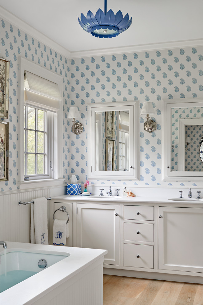 Photo of a coastal bathroom in Boston with recessed-panel cabinets, white cabinets, blue walls, light hardwood flooring, white worktops, double sinks, a built in vanity unit and wallpapered walls.