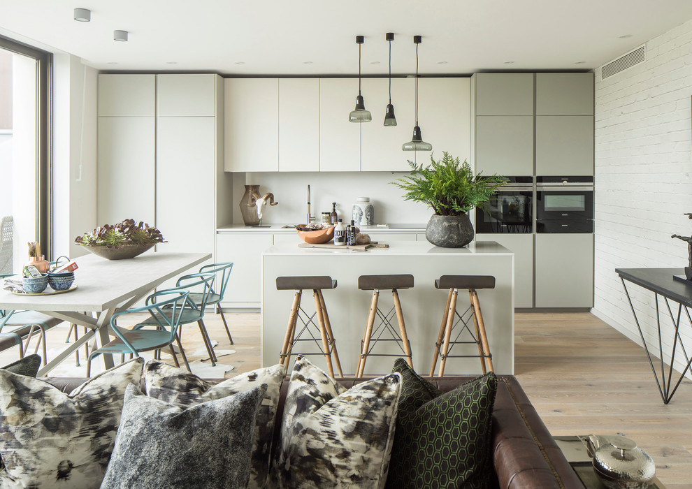 Design ideas for a contemporary open plan kitchen in London with flat-panel cabinets, white cabinets, white splashback, black appliances, light hardwood floors, with island and white benchtop.