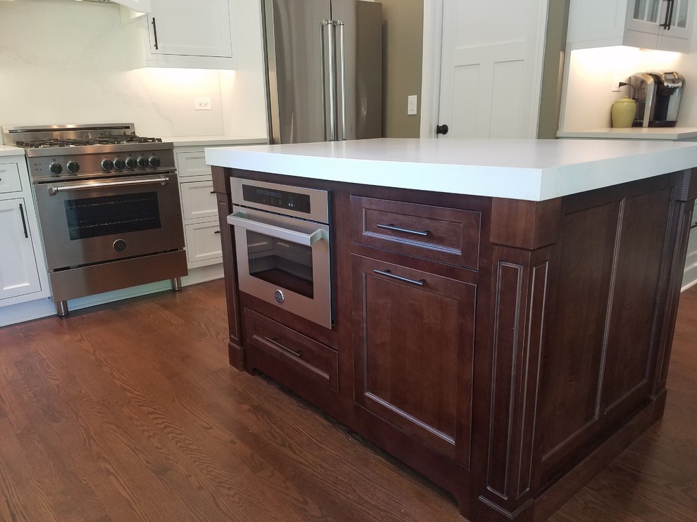 Inspiration for a mid-sized transitional l-shaped medium tone wood floor and brown floor eat-in kitchen remodel in Chicago with a farmhouse sink, recessed-panel cabinets, white cabinets, quartz countertops, white backsplash, stainless steel appliances and an island