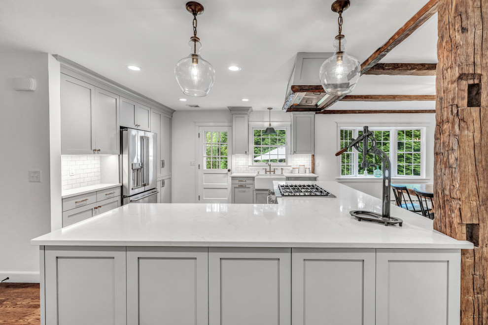 Inspiration for a large country u-shaped eat-in kitchen in New York with a farmhouse sink, shaker cabinets, grey cabinets, quartz benchtops, white splashback, subway tile splashback, stainless steel appliances, medium hardwood floors, with island, brown floor, white benchtop and exposed beam.