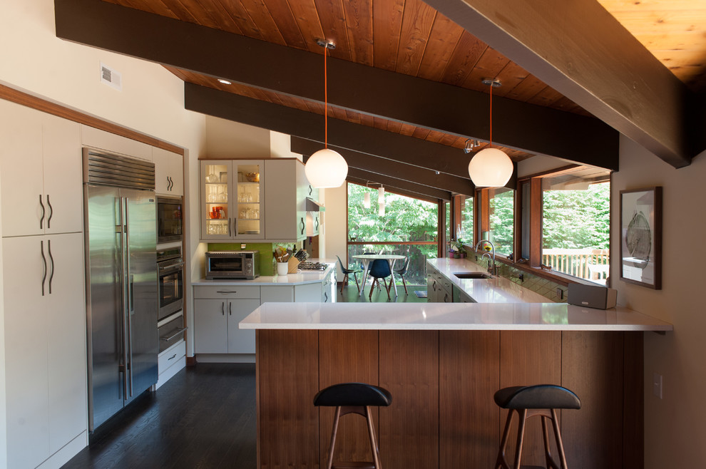 This is an example of a large midcentury l-shaped eat-in kitchen in Wilmington with an undermount sink, flat-panel cabinets, quartz benchtops, green splashback, stainless steel appliances, dark hardwood floors, a peninsula, brown floor and white benchtop.