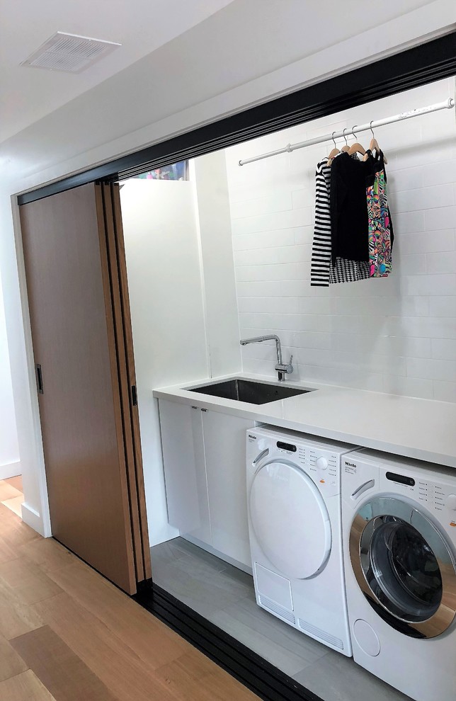 This is an example of a small modern single-wall laundry cupboard in Toronto with an undermount sink, quartz benchtops, white walls, a side-by-side washer and dryer, white benchtop, open cabinets, white cabinets, light hardwood floors and brown floor.
