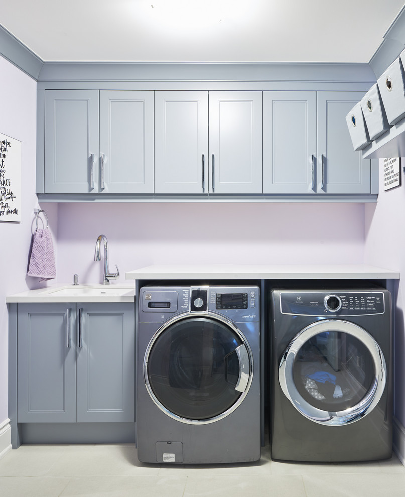 This is an example of a mid-sized contemporary single-wall utility room in Toronto with an undermount sink, shaker cabinets, grey cabinets, quartz benchtops, purple walls, porcelain floors, a side-by-side washer and dryer, white floor and white benchtop.