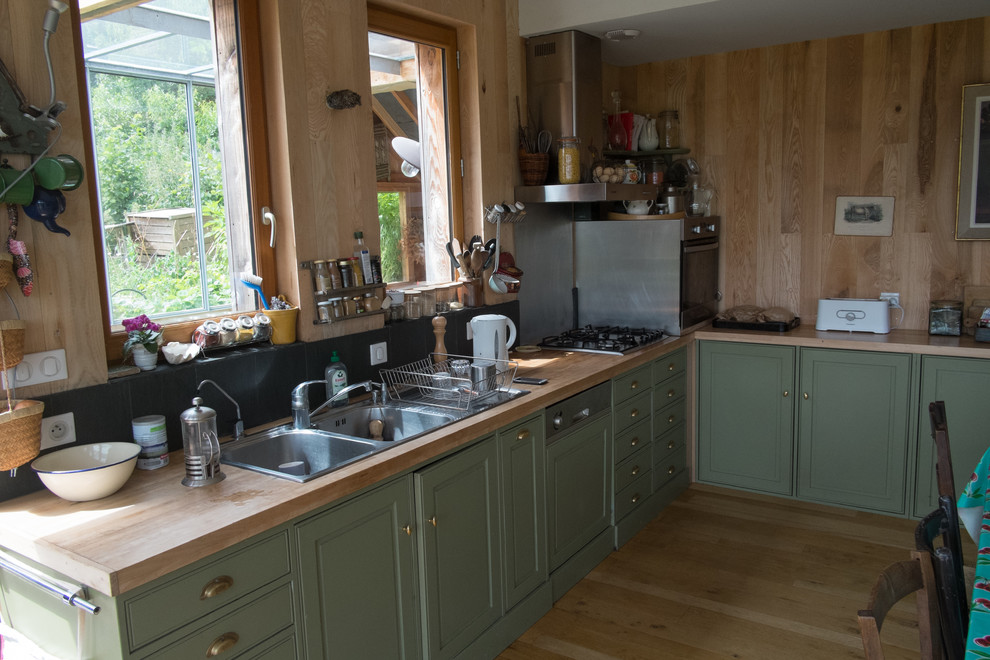 This is an example of a mid-sized country l-shaped open plan kitchen in Paris with a double-bowl sink, green cabinets, wood benchtops, black splashback, porcelain splashback, coloured appliances, light hardwood floors, no island, brown floor and brown benchtop.