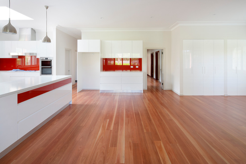 Design ideas for a large contemporary u-shaped eat-in kitchen in Sydney with a drop-in sink, flat-panel cabinets, white cabinets, quartz benchtops, red splashback, glass sheet splashback, stainless steel appliances, bamboo floors, a peninsula, brown floor and white benchtop.
