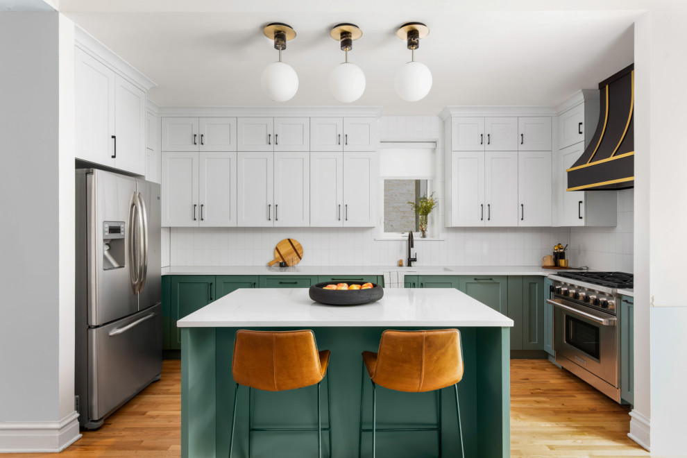 This is an example of a mid-sized transitional u-shaped eat-in kitchen in Chicago with a drop-in sink, shaker cabinets, green cabinets, quartz benchtops, white splashback, ceramic splashback, stainless steel appliances, medium hardwood floors, with island, brown floor and white benchtop.