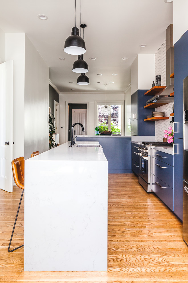 Photo of a mid-sized transitional l-shaped kitchen in San Francisco with a farmhouse sink, flat-panel cabinets, blue cabinets, quartz benchtops, white splashback, ceramic splashback, medium hardwood floors, with island, grey benchtop, stainless steel appliances and brown floor.