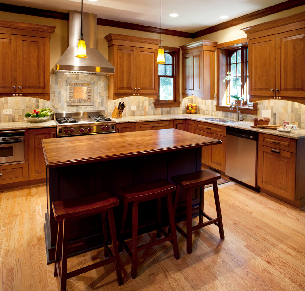This is an example of a mid-sized arts and crafts l-shaped eat-in kitchen in Chicago with with island, granite benchtops, stainless steel appliances, a double-bowl sink, raised-panel cabinets, medium wood cabinets, multi-coloured splashback, stone tile splashback and light hardwood floors.