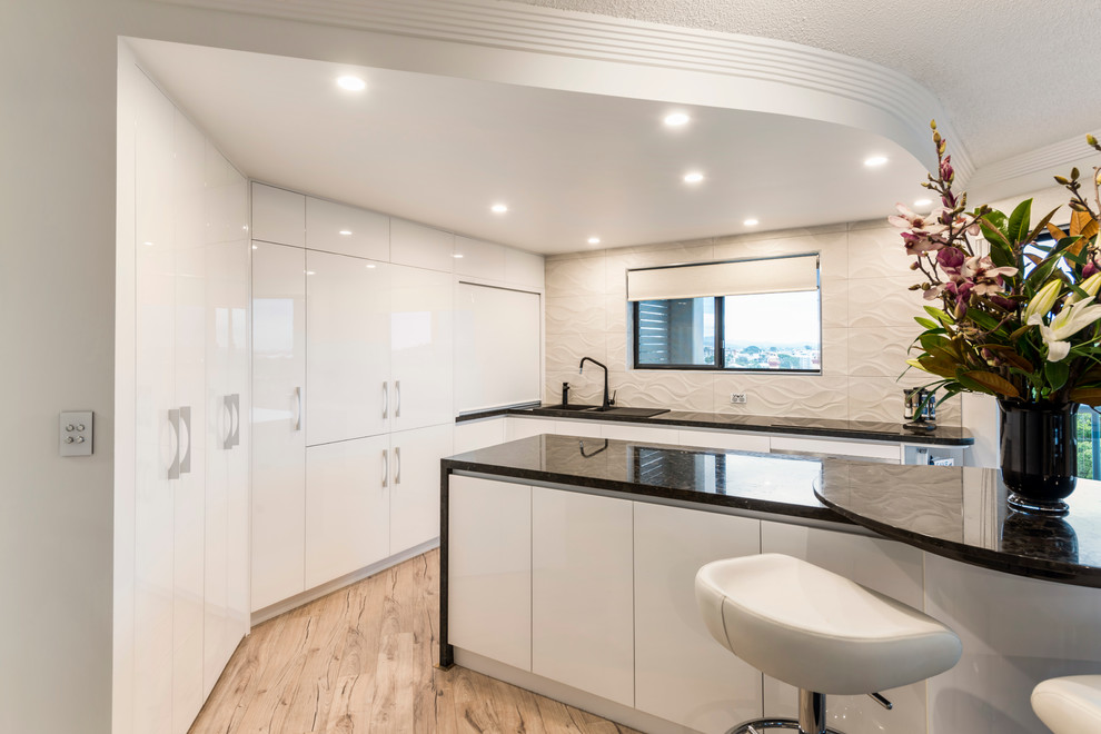 Design ideas for a mid-sized contemporary l-shaped eat-in kitchen in Brisbane with a drop-in sink, flat-panel cabinets, white cabinets, granite benchtops, beige splashback, cement tile splashback, stainless steel appliances, light hardwood floors and with island.