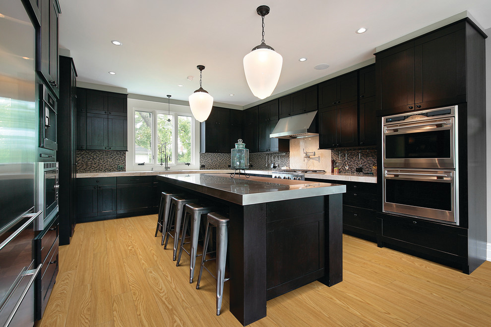 Large transitional u-shaped light wood floor enclosed kitchen photo with an undermount sink, shaker cabinets, dark wood cabinets, stainless steel countertops, multicolored backsplash, mosaic tile backsplash, stainless steel appliances and an island