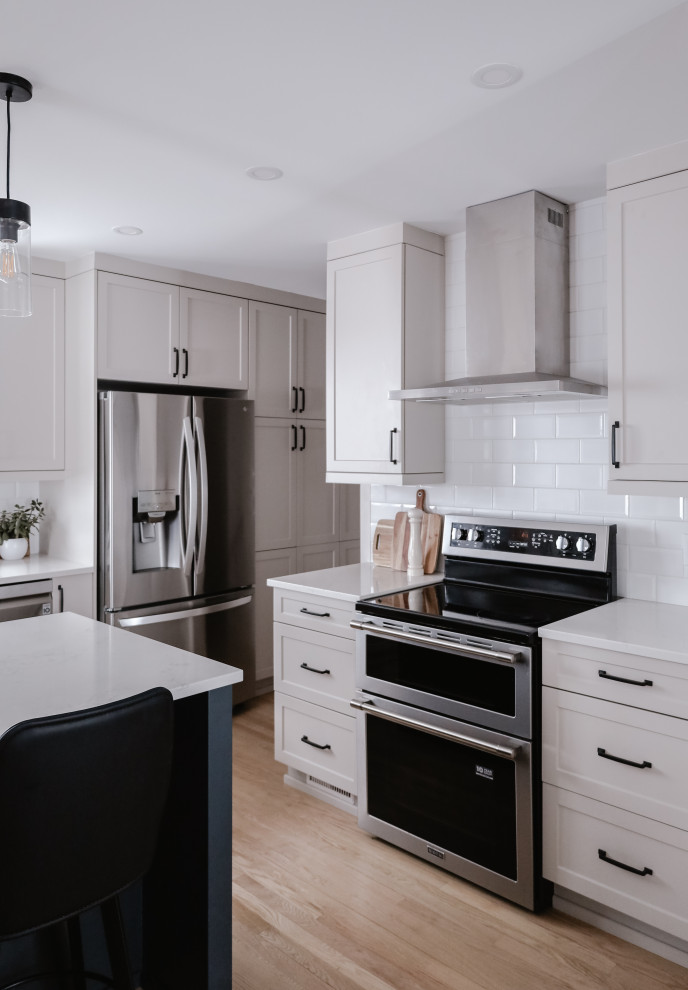 Mid-sized transitional l-shaped light wood floor and brown floor eat-in kitchen photo with an undermount sink, shaker cabinets, beige cabinets, quartz countertops, white backsplash, subway tile backsplash, stainless steel appliances, an island and white countertops