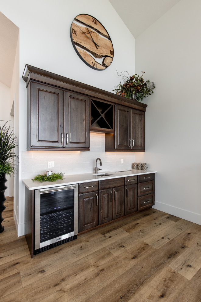Inspiration for a mid-sized country single-wall wet bar in Calgary with an undermount sink, raised-panel cabinets, distressed cabinets, quartz benchtops, white splashback, ceramic splashback, medium hardwood floors, brown floor and white benchtop.