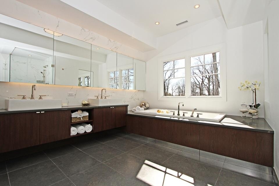 This is an example of a large modern master bathroom in New York with flat-panel cabinets, medium wood cabinets, a drop-in tub, gray tile, white tile, stone slab, white walls, porcelain floors, a vessel sink and grey floor.