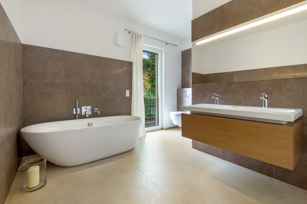 Photo of a mid-sized modern master bathroom in Cologne with flat-panel cabinets, medium wood cabinets, a freestanding tub, a one-piece toilet, beige tile, stone tile, limestone floors, brown walls and an integrated sink.