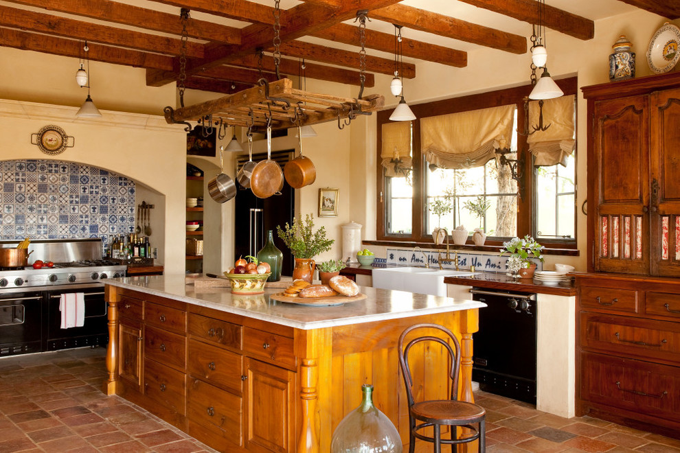 Kitchen - mediterranean l-shaped terra-cotta tile kitchen idea in Orange County with a farmhouse sink, raised-panel cabinets, medium tone wood cabinets, multicolored backsplash, black appliances and an island