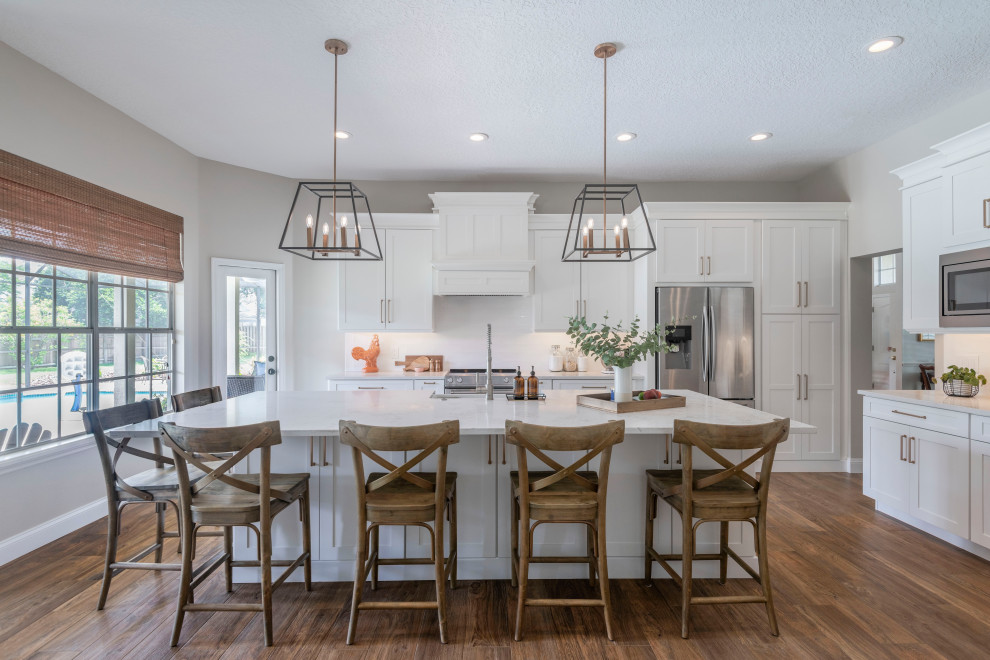 Inspiration for a l-shaped kitchen in Orlando with a farmhouse sink, shaker cabinets, white cabinets, white splashback, stainless steel appliances, dark hardwood floors, with island, brown floor and white benchtop.