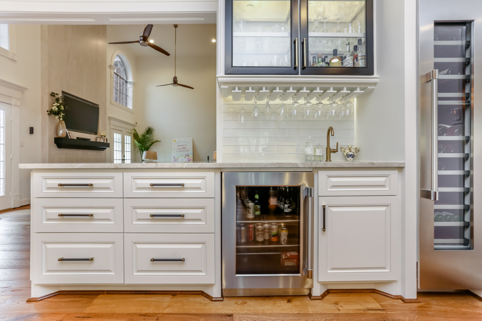 Photo of a mid-sized transitional single-wall wet bar in Houston with an undermount sink, glass-front cabinets, white cabinets, granite benchtops, white splashback, glass tile splashback, medium hardwood floors, brown floor and white benchtop.