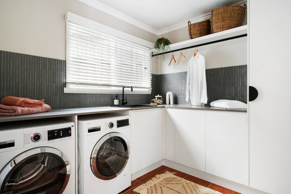 Inspiration for a mid-sized contemporary l-shaped utility room in Melbourne with a drop-in sink, flat-panel cabinets, white cabinets, quartz benchtops, grey splashback, matchstick tile splashback, white walls, porcelain floors, a side-by-side washer and dryer, orange floor and grey benchtop.