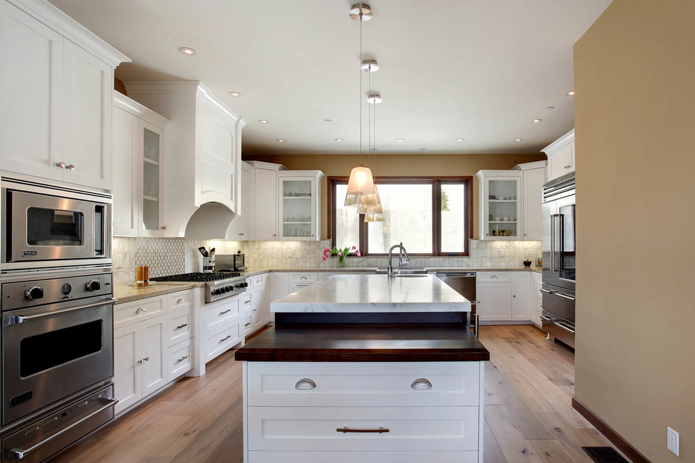 Large transitional u-shaped separate kitchen in Orange County with a farmhouse sink, recessed-panel cabinets, white cabinets, marble benchtops, white splashback, marble splashback, stainless steel appliances, medium hardwood floors, with island and brown floor.