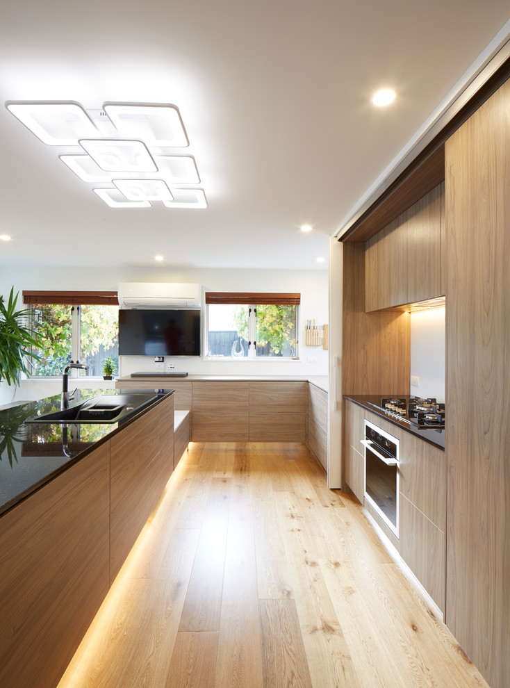 Expansive contemporary u-shaped eat-in kitchen in Christchurch with a drop-in sink, glass-front cabinets, white cabinets, granite benchtops, white splashback, glass sheet splashback, black appliances, plywood floors and a peninsula.