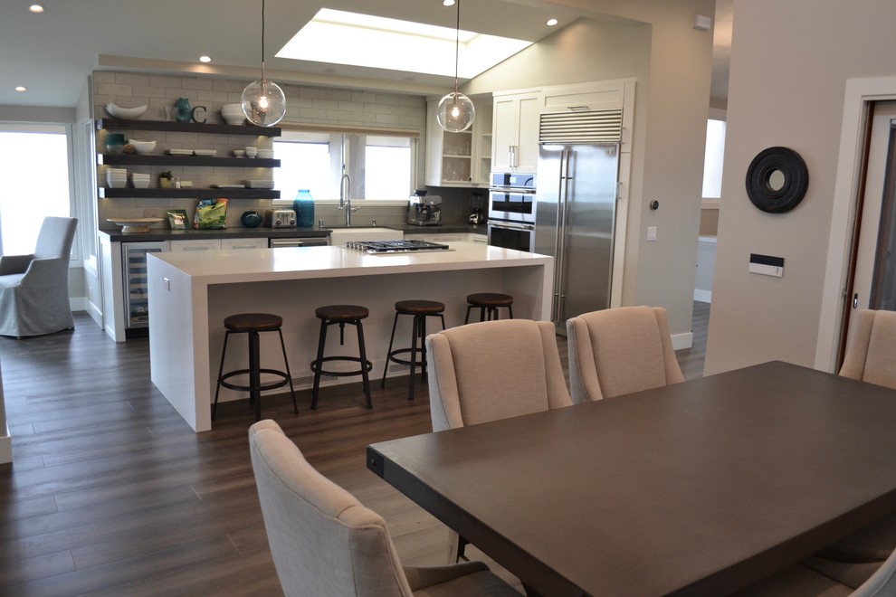 Small contemporary single-wall eat-in kitchen in Orange County with a farmhouse sink, shaker cabinets, white cabinets, quartz benchtops, grey splashback, glass tile splashback, stainless steel appliances, medium hardwood floors, with island and brown floor.