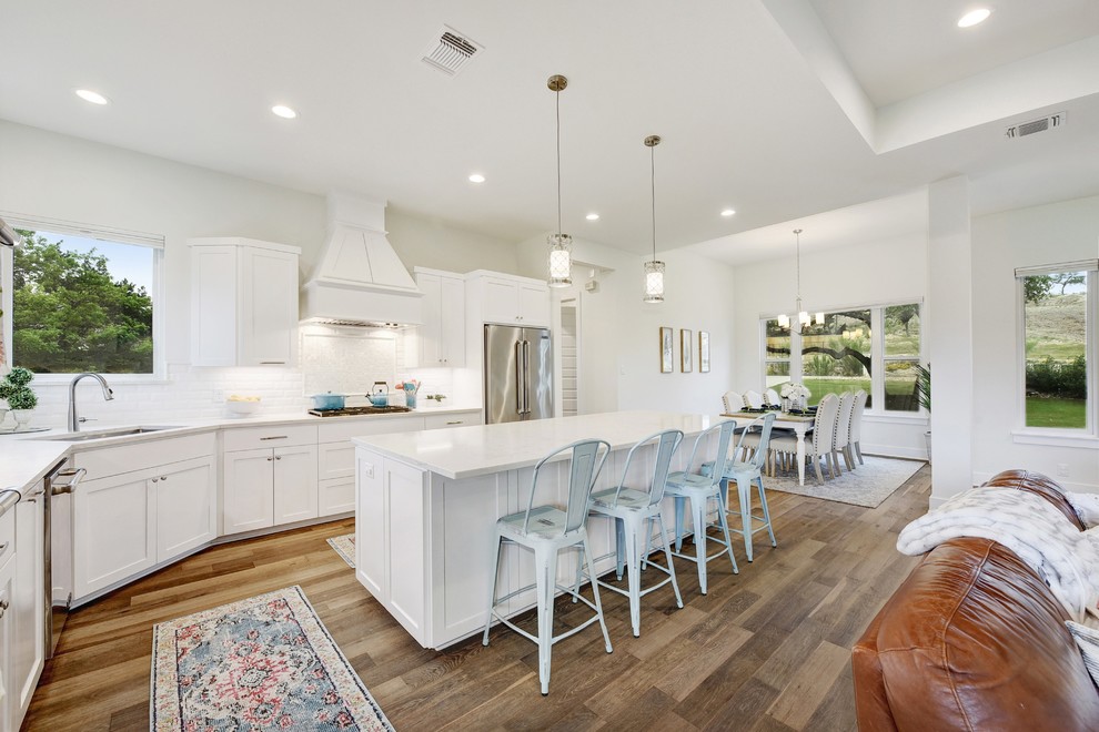 Example of a transitional l-shaped medium tone wood floor and brown floor eat-in kitchen design in Austin with an undermount sink, shaker cabinets, white cabinets, white backsplash, subway tile backsplash, stainless steel appliances, an island and white countertops