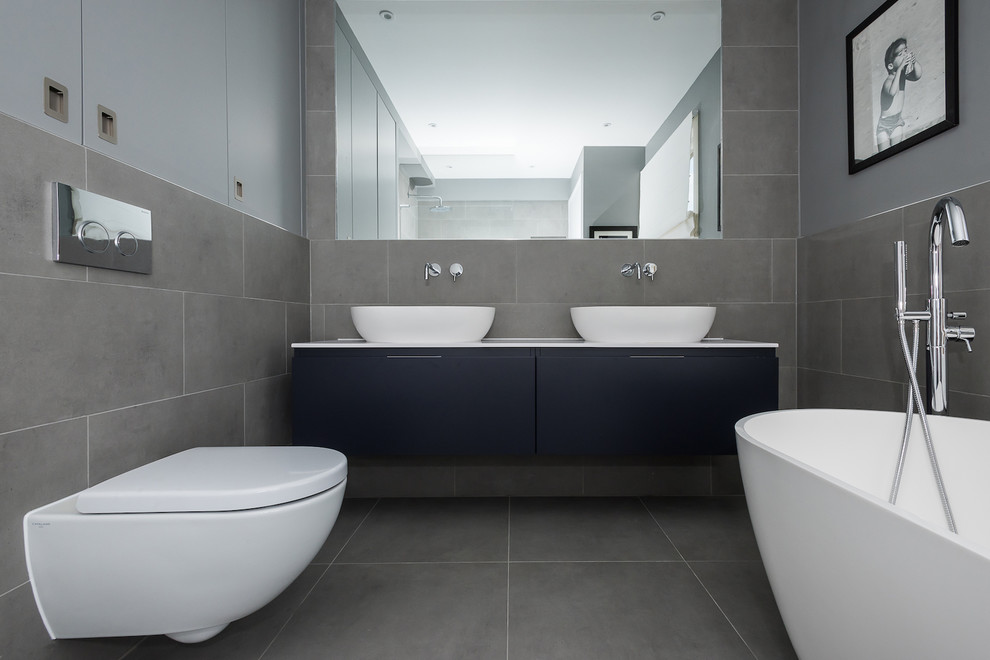 This is an example of a large modern kids bathroom in London with blue cabinets, a freestanding tub, a shower/bathtub combo, a wall-mount toilet, gray tile, ceramic tile, green walls and ceramic floors.
