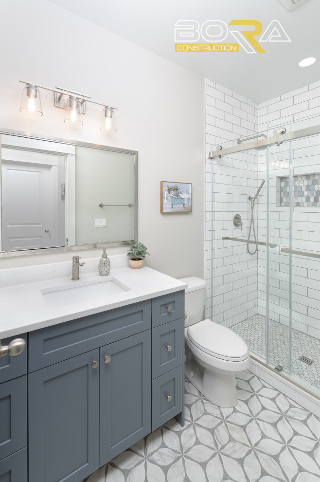 На фото: маленькая ванная комната в стиле неоклассика (современная классика) с фасадами в стиле шейкер, синими фасадами, душем над ванной, раздельным унитазом, белой плиткой, керамической плиткой, белыми стенами, душевой кабиной, врезной раковиной, столешницей из искусственного кварца, душем с раздвижными дверями, белой столешницей, тумбой под одну раковину и напольной тумбой для на участке и в саду