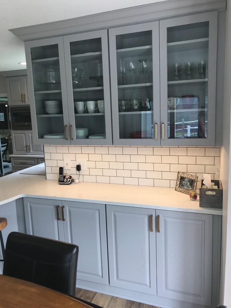 Mid-sized transitional u-shaped vinyl floor and brown floor enclosed kitchen photo with an undermount sink, raised-panel cabinets, gray cabinets, quartz countertops, white backsplash, ceramic backsplash, a peninsula and white countertops