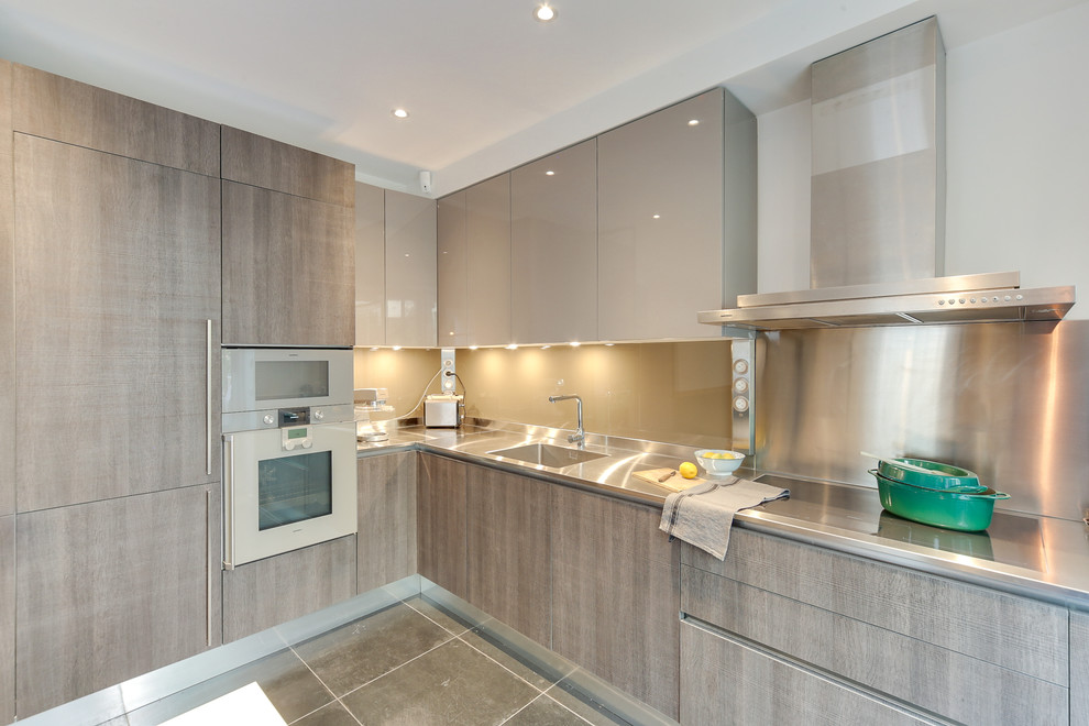 Large contemporary l-shaped eat-in kitchen in Paris with glass sheet splashback, stainless steel appliances, ceramic floors, no island, an integrated sink, flat-panel cabinets, grey cabinets, stainless steel benchtops, grey splashback, grey floor and grey benchtop.