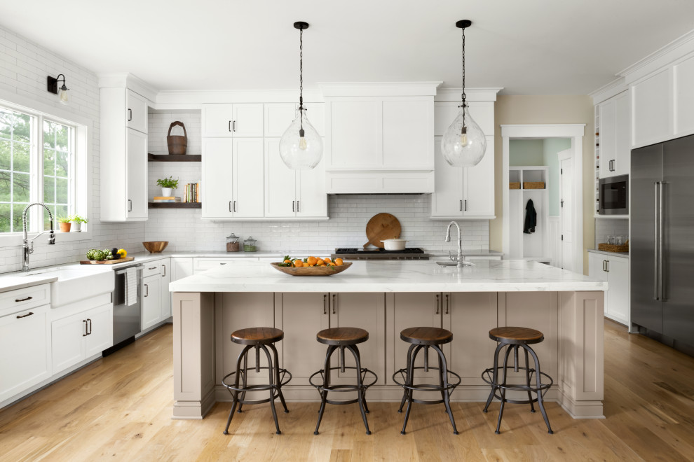 Photo of a transitional kitchen in DC Metro with a farmhouse sink, shaker cabinets, white cabinets, quartz benchtops, white splashback, ceramic splashback, stainless steel appliances, light hardwood floors, with island and white benchtop.