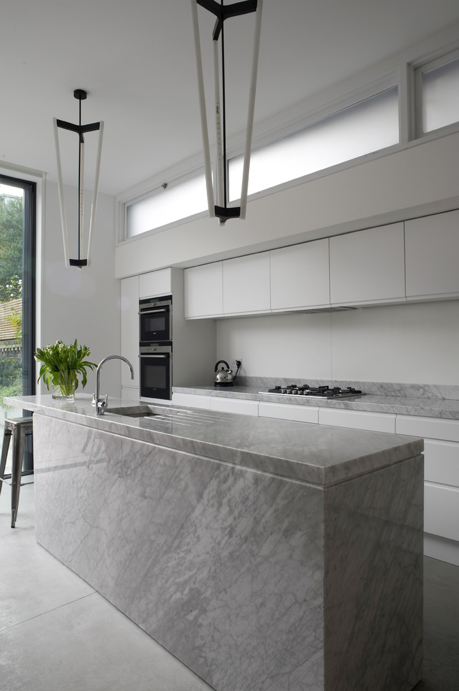 Mid-sized modern single-wall eat-in kitchen in London with an undermount sink, flat-panel cabinets, marble benchtops, white splashback, glass sheet splashback, stainless steel appliances, concrete floors, with island and white cabinets.